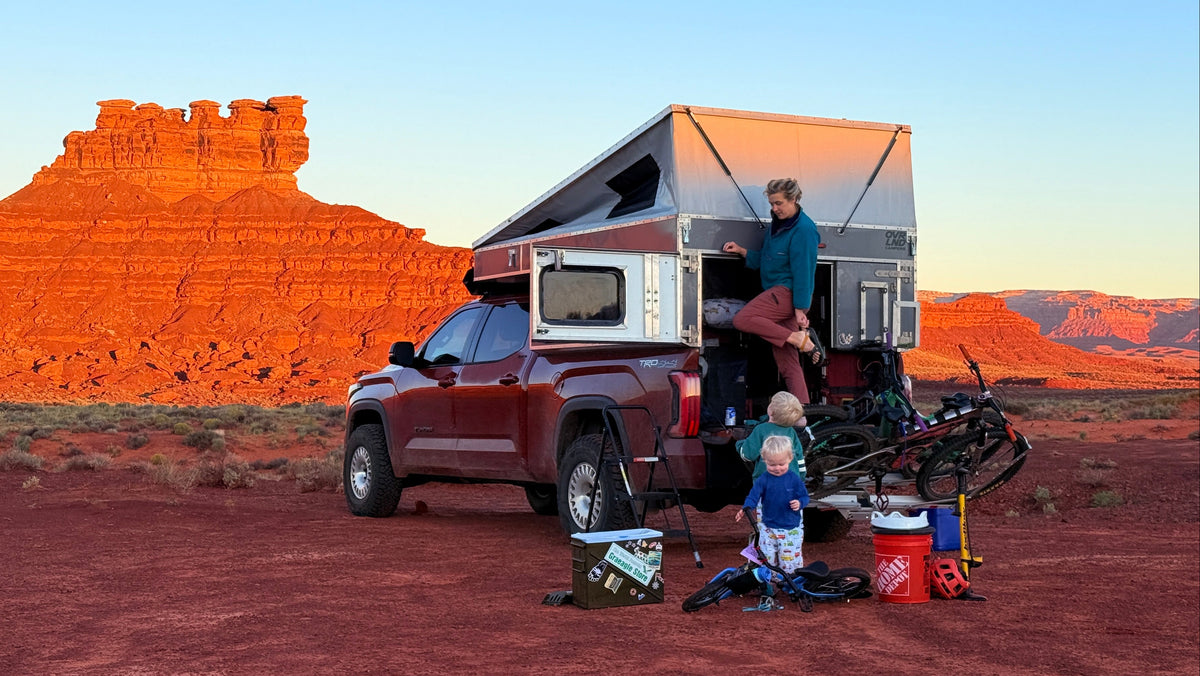 A family camping setup in a desert landscape with red rock buttes at sunset. A dark red pickup truck has a large pop-up camper attached, with a man standing at the back door and a child sitting on the ground nearby.